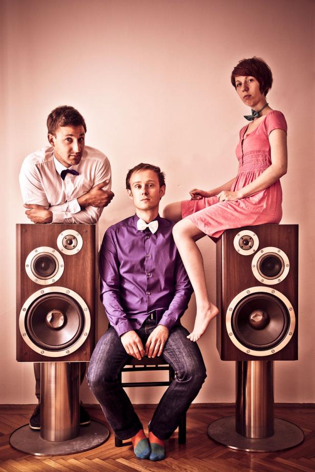 Drei Personen posieren mit Fliege auf oder an großen Lautsprecherboxen.