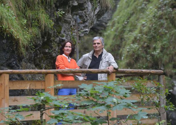 Ein Paar steht auf einer Holzbrücke in einer grünen, bewaldeten Umgebung.