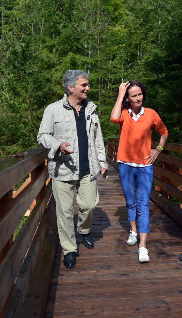 Ein Mann und eine Frau spazieren auf einer Holzbrücke vor einem Wald.