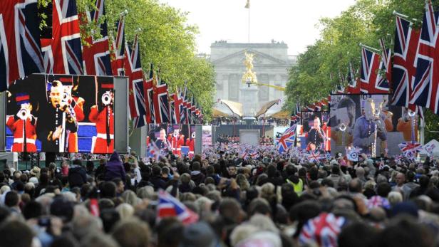 Eine Menschenmenge feiert vor dem Buckingham Palace, geschmückt mit Union-Jack-Flaggen.