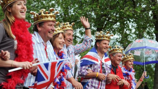 Eine Gruppe Menschen mit Kronen und Union-Jack-Flaggen feiert fröhlich.