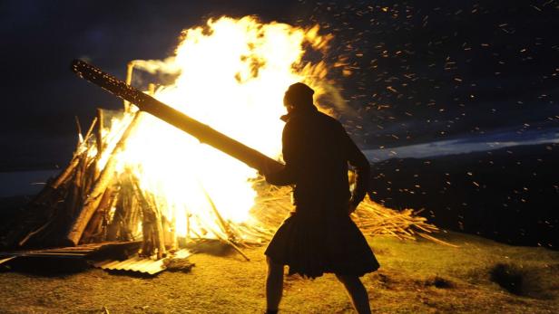Eine Person in einem Kilt steht vor einem großen Lagerfeuer und hält einen brennenden Baumstamm.
