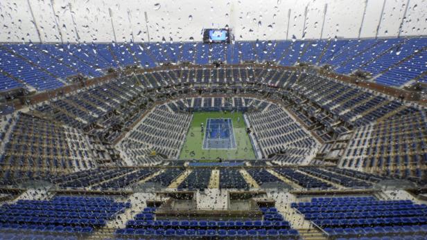 Blick durch eine regennasse Scheibe auf ein leeres Tennisstadion mit blauen Sitzen.
