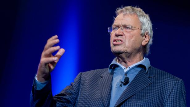 Ein älterer Mann mit Brille gestikuliert vor einem blauen Hintergrund.