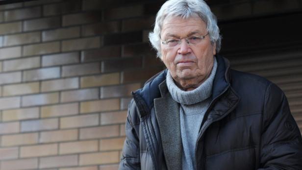 Ein älterer Mann mit Brille und grauen Haaren steht vor einer Backsteinmauer.