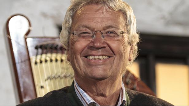 Ein lächelnder älterer Mann mit Brille vor einer Zither.