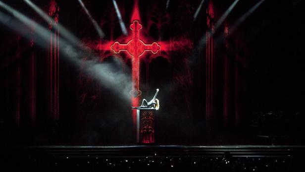 Madonna liegt auf der Bühne vor einem großen roten Kreuz.