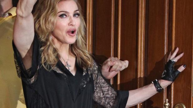 Madonna posiert vor einer Holztür mit einem schwarzen Kleid und einem Kreuzanhänger.