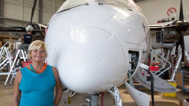 Eine Frau in einem blauen Kleid steht vor dem Bug eines kleinen Flugzeugs in einer Werfthalle.