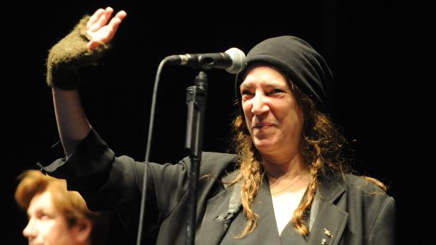 Patti Smith tritt in einer schwarzen Jacke und Mütze vor einem Mikrofon auf.