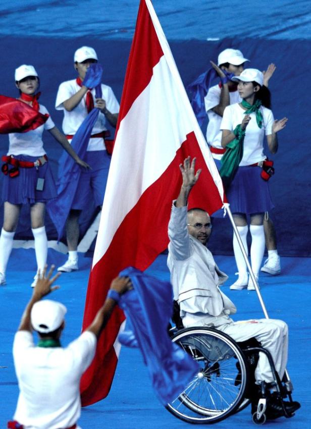 Ein Mann im Rollstuhl schwenkt eine rot-weiße Flagge, umgeben von applaudierenden Personen.