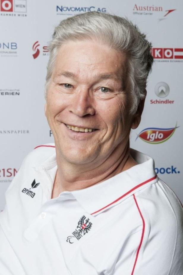 Ein lächelnder Mann mit grauen Haaren trägt ein weißes Poloshirt mit dem österreichischen Wappen.