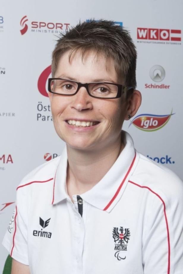 Eine lächelnde Frau mit Brille und Sportbekleidung vor einem Sponsorenhintergrund.