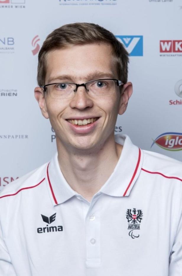 Ein lächelnder Mann mit Brille und einem Poloshirt mit dem Aufdruck „Austria“.
