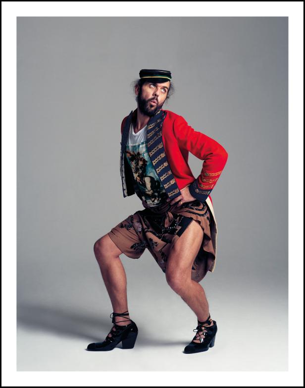 Ein Mann in einer roten Jacke, Shorts und hochhackigen Schuhen posiert vor grauem Hintergrund.