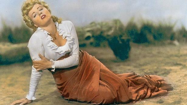 Eine Frau mit blonden Locken liegt in einem langen, orangefarbenen Rock in der Natur.