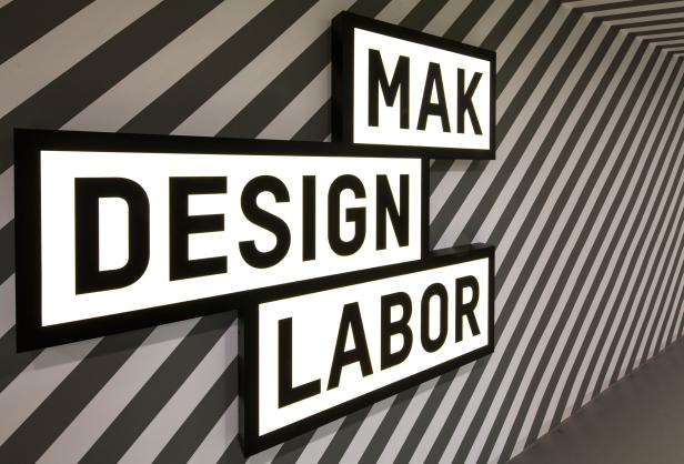Leuchtschilder mit der Aufschrift „MAK Design Labor“ an einer gestreiften Wand.