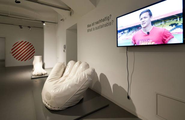 In einer Ausstellung sind eine übergroße Hand und ein Fuß ausgestellt, während ein Mann im Fernsehen über Nachhaltigkeit spricht.