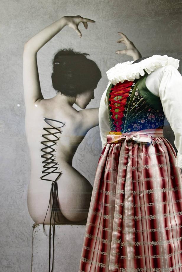 Eine Schaufensterpuppe mit einem Dirndl vor einem Aktfoto mit Korsett-Piercings.