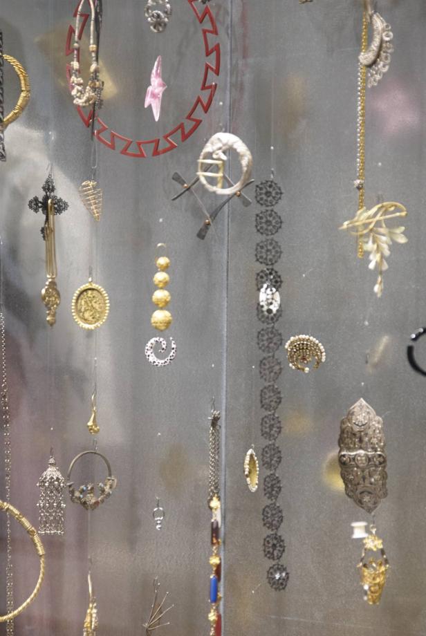 Verschiedene Schmuckstücke hängen an Fäden vor einem grauen Hintergrund.