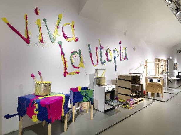 Eine Kunstinstallation mit dem Schriftzug „Viva la Utopia“ an einer Wand und bemalten Möbeln.