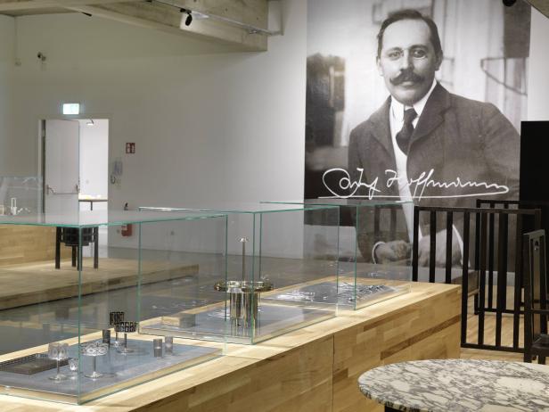 Ausstellungsraum mit Glasvitrinen, die Designobjekte und ein Porträt von Josef Hoffmann zeigen.