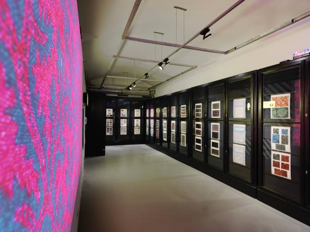 Ein Ausstellungsraum mit Mustern in Vitrinen und einer farbenfrohen Wand im Vordergrund.