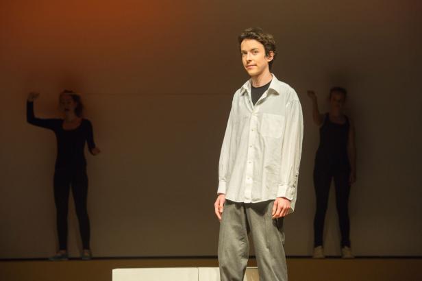 Ein junger Mann in einem weißen Hemd steht auf einer Bühne, im Hintergrund zwei Tänzerinnen.