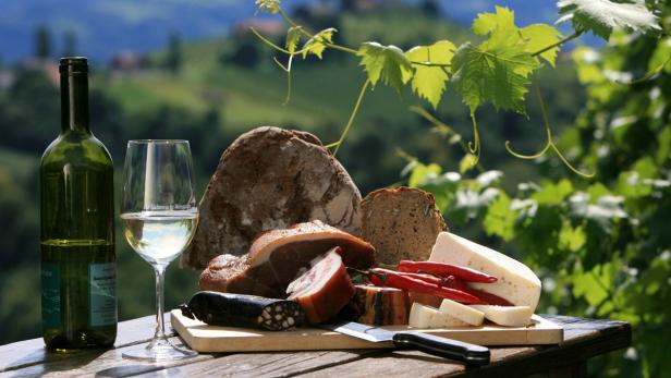 Eine Flasche Wein, Brot, Wurst, Käse und Paprika auf einem Holztisch im Freien.