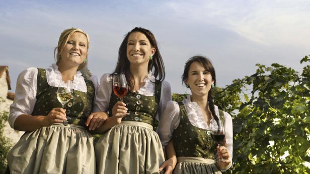 Drei junge Frauen in Dirndln halten Weingläser in einem Weinberg.