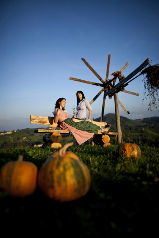 Zwei Frauen sitzen auf einer Bank vor einer Windmühle, im Vordergrund Kürbisse.