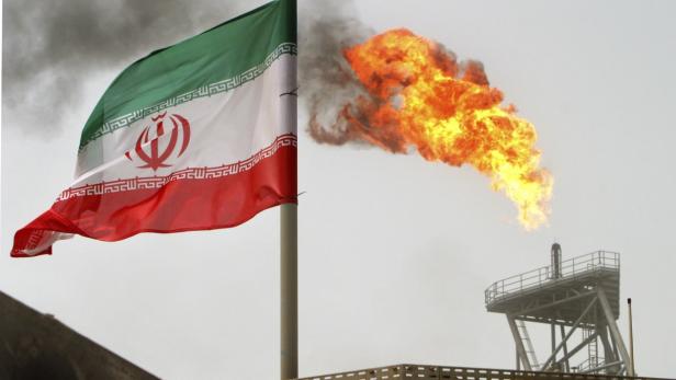 Die iranische Flagge weht vor einer Ölraffinerie, aus der Flammen schlagen.