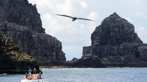 Ein Boot mit Personen fährt zwischen zwei felsigen Inseln hindurch, während ein Vogel darüber fliegt.