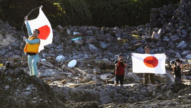Mehrere Personen halten japanische Flaggen an einem felsigen Strand.