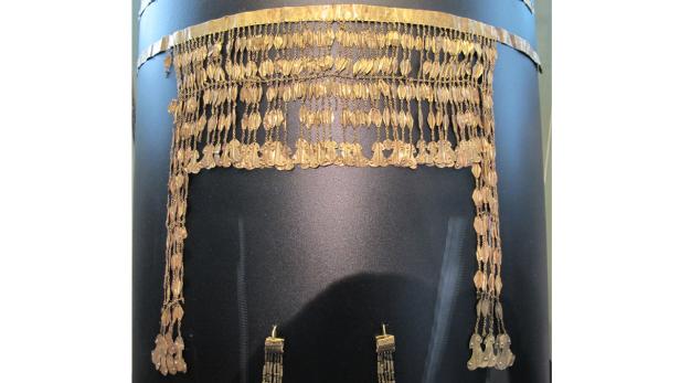 Ein goldenes Stirnband mit hängenden Blättchen und Fransen wird auf einem schwarzen Sockel präsentiert.