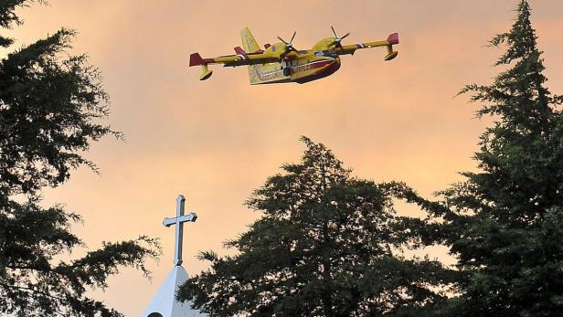 Ein gelbes Löschflugzeug fliegt über eine Kirche mit Kreuz im Gegenlicht.