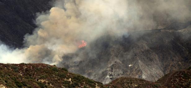 Ein Waldbrand wütet in den Bergen, dichter Rauch steigt auf. Ein Hubschrauber ist im Einsatz.