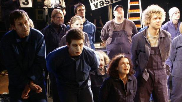 Eine Gruppe von Menschen in blauen Overalls steht auf einer Bühne.