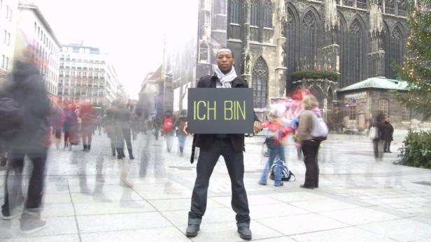 Ein Mann hält ein Schild mit der Aufschrift „Ich bin“ in einer belebten Straße in Wien.