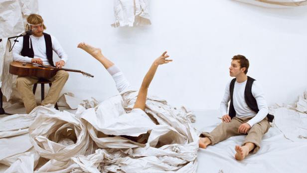 Ein Mann spielt Gitarre, während ein anderer Mann zusieht, wie eine Person in Papier gehüllt ist.