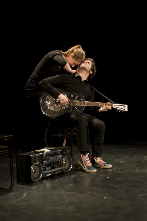 Eine Frau beugt sich über einen Gitarre spielenden Mann, neben dem ein Ghettoblaster steht.