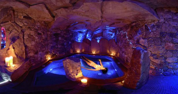 Eine Frau entspannt in einem Whirlpool in einer Grotte, beleuchtet mit Kerzen und blauem Licht.