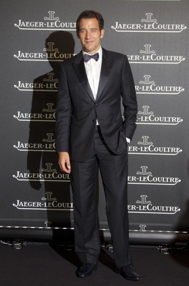 Clive Owen in einem dunklen Anzug mit Fliege vor einer Wand mit Jaeger-LeCoultre Logos.