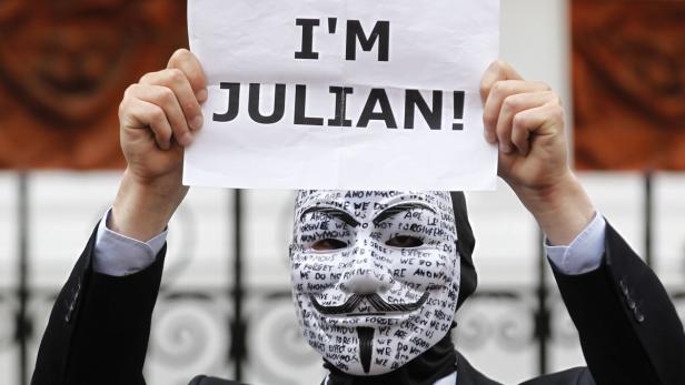 Eine Person mit einer Guy-Fawkes-Maske hält ein Schild mit der Aufschrift „I'M JULIAN!“ hoch.
