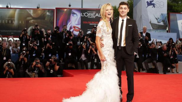 Zac Efron und eine Frau in einem weißen Kleid posieren auf einem roten Teppich vor Fotografen.