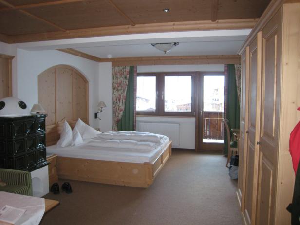 Ein helles Schlafzimmer mit Holzbalkendecke und einem Kachelofen neben dem Bett.