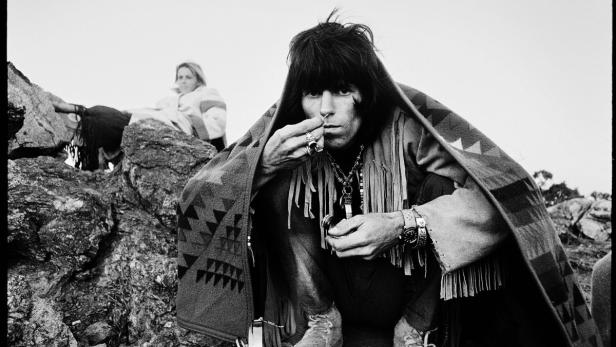 Keith Richards, gehüllt in eine Decke, hockt auf einem Felsen.