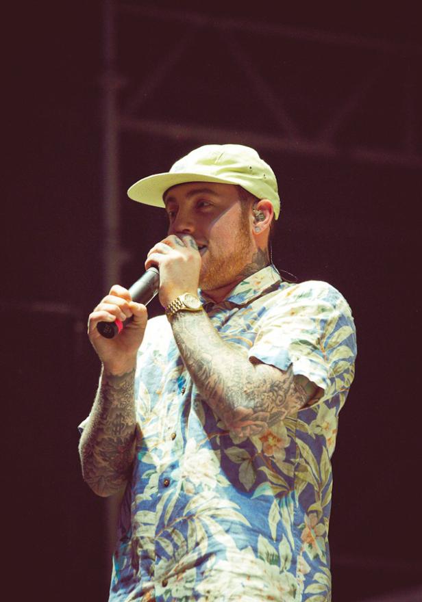 Mac Miller tritt mit Tätowierungen und einem floralen Hemd auf einer Bühne auf.