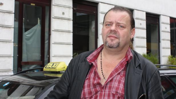 Ein Mann mit Jacke und kariertem Hemd steht vor einem Taxi.