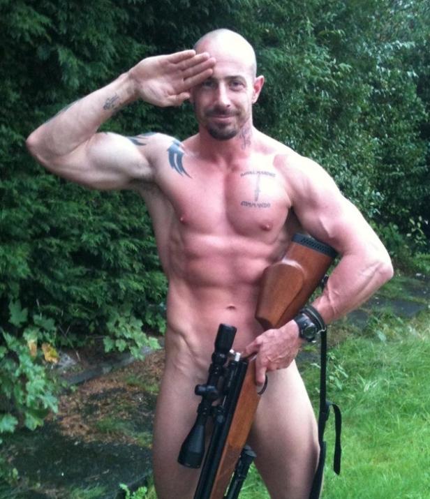 Ein muskulöser, nackter Mann mit Tätowierungen salutiert und hält ein Gewehr.